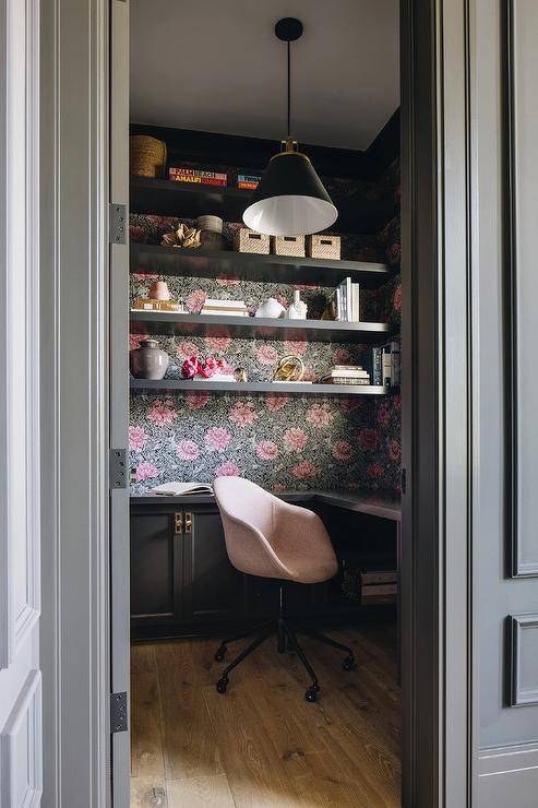 Black floating shelves are mounted against a wall clad in pink and black floral wallpaper and over a black l-shaped desk paired with a pink chair lit by a Rejuvenation Butte Cone Pendant.