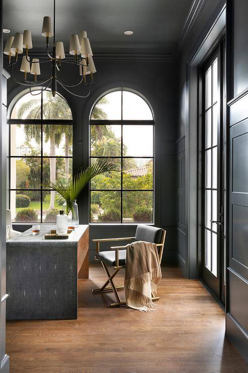 Dark gray walls finished with dark gray wainscoting framed arch windows allowing natural light to stream into a home office boasting a gold and black waterfall desk lit by a Farlane chandelier hung from a dark gray ceiling lined with dark gray crown moldings.
