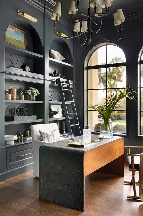 Chic dark gray office features a gold and black waterfall desk paired with a white chair and placed in front of arched dark gray built-in shelves lit by brass picture lights and matched with a dark gray ladder on rails. The room is illuminated by a Farlane Chandelier and natural light streaming in front arch windows framed by a dark gray wall.