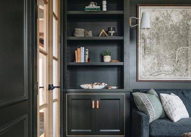 Styled built-in olive green living room shelves are mounted over cabinets accented with leather tab pulls. While a map art piece is mounted above a black skirted sofa positioned on layered rugs.