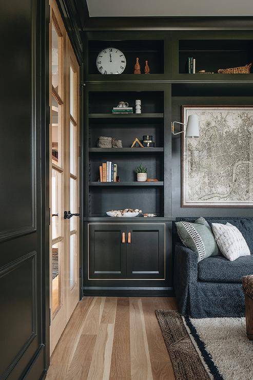 Styled built-in olive green living room shelves are mounted over cabinets accented with leather tab pulls. While a map art piece is mounted above a black skirted sofa positioned on layered rugs.