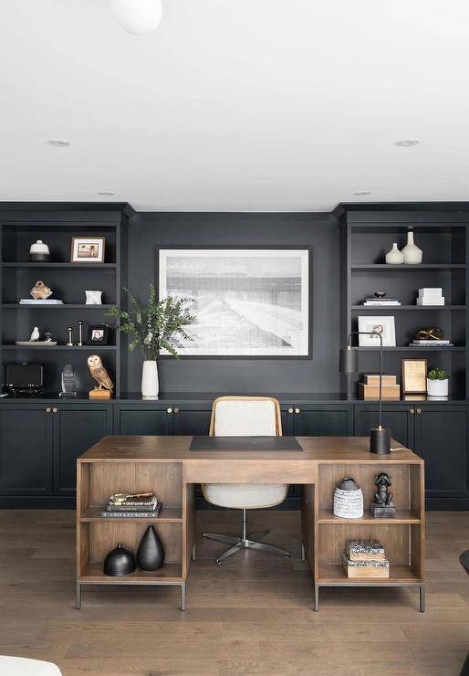 Chic black and brown home office boasts a brown wooden desk matched with a white and brown desk chair placed in front of black built-in cabinets fixed under black shelves flanking a black and white photograph.