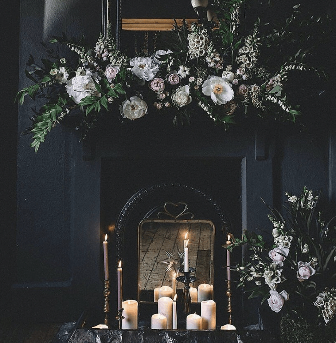 dark and moody fireplace with fl، arrangements and white lit candles inside completely black