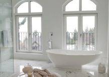 Luxurious master bathroom with crystal chandelier over round white bathroom ottoman over white and gray marble tile floor. Master bathroom features freestanding bathtub placed in front of French doors leading to Juliet balconies and glass walk-in shower.