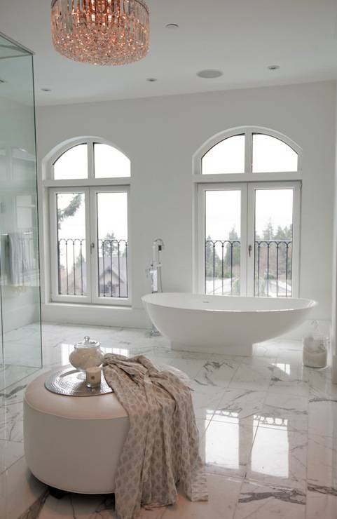 Luxurious master bathroom with crystal chandelier over round white bathroom ottoman over white and gray marble tile floor. Master bathroom features freestanding bathtub placed in front of French doors leading to Juliet balconies and glass walk-in shower.