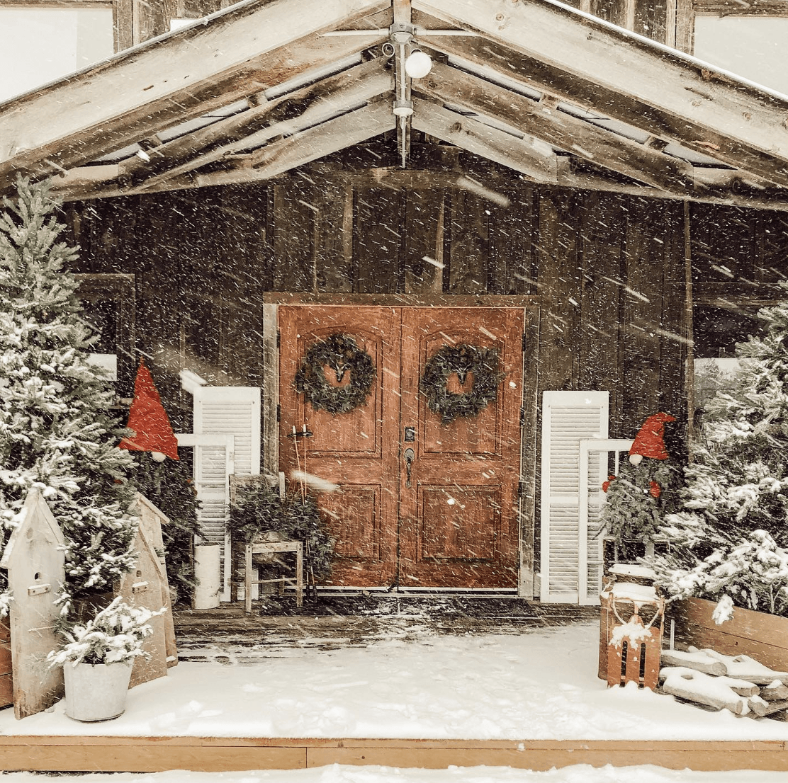 old antique barn in country snowing with double wreaths on red doors christmas trees covered in snow old shutters