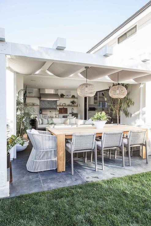Gray rope dining chairs surround a teak outdoor dining table placed beneath a pergola.