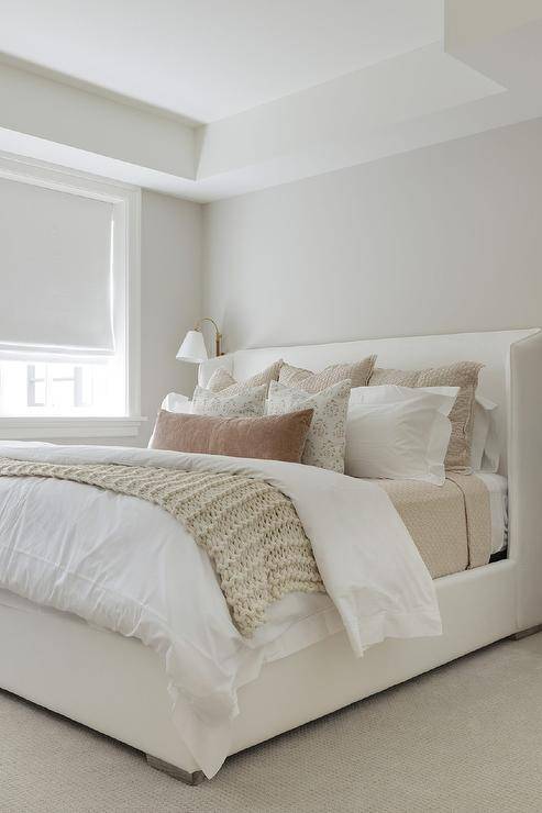 A white upholstered wingback bed is complemented with tan and brown layered pillows placed on white and tan bedding.