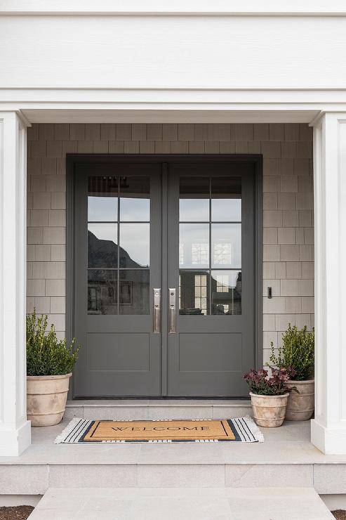 Styled front porch features layered welcome mats in front of gray and glass front door finished with potted plants.