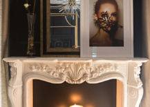 Master bedroom fireplace features an ornate carved marble fireplace mantel filled with lit candles and topped with a vase of spring blossoms, graffitied photo portrait and black and gold mirror framed by black walls.