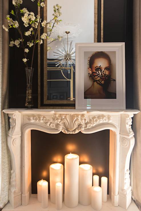 Master bedroom fireplace features an ornate carved marble fireplace mantel filled with lit candles and topped with a vase of spring blossoms, graffitied photo portrait and black and gold mirror framed by black walls.