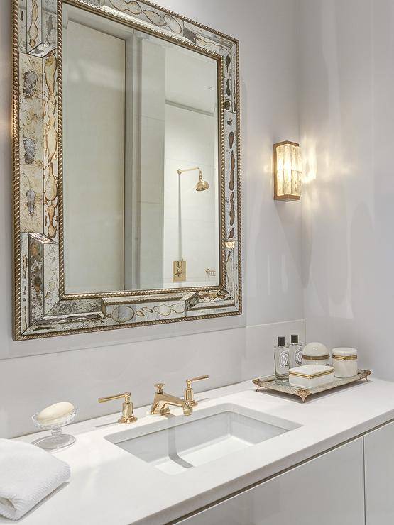 White and gold bathroom features an antiqued mirror beveled mirror over a white washstand topped with white quartz fitted with a sink and gold faucet next to a an antiqued mirrored footed tray.