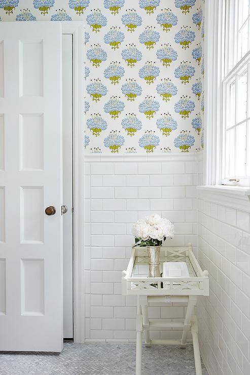 Lovely bathroom features upper walls clad in blue and green floral wallpaper and lower walls clad in white subway tiles lined with a white bamboo tray table atop a marble herringbone floor next to a multi paneled door.