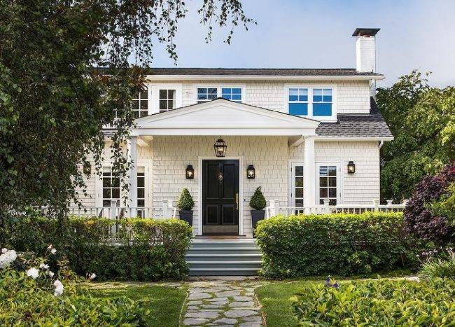 White cottage home with white shingles and a gray roof contrasted with a black front door and black outdoor carriage lantern sconces. A covered porch displays doric columns for a classic finish contributing to a stunning curb appeal.