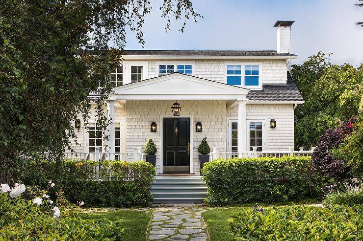 White cottage home with white shingles and a gray roof contrasted with a black front door and black outdoor carriage lantern sconces. A covered porch displays doric columns for a classic finish contributing to a stunning curb appeal.