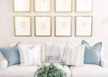 Gold framed gallery wall on a white wall adds a sophisticated appeal above a white sofa with Greek key trim and blue pillows. A light gray oak coffee table on a tan and white geometric rug completes the living space with a cozy finish.