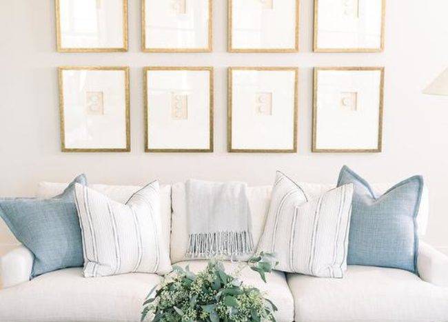 Gold framed gallery wall on a white wall adds a sophisticated appeal above a white sofa with Greek key trim and blue pillows. A light gray oak coffee table on a tan and white geometric rug completes the living space with a cozy finish.