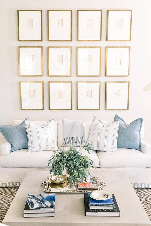 Gold framed gallery wall on a white wall adds a sophisticated appeal above a white sofa with Greek key trim and blue pillows. A light gray oak coffee table on a tan and white geometric rug completes the living space with a cozy finish.