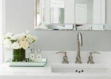 White and gray bathroom features a gray wall lined with a beveled vanity mirror over a white lacquered washstand fitted with dresser like drawers adorned with ring pulls topped with white quartz framing a white porcelain sink and nickel gooseneck faucet alongside a mirrored beveled tray filled with white roses and Jo Malone Fragrances.