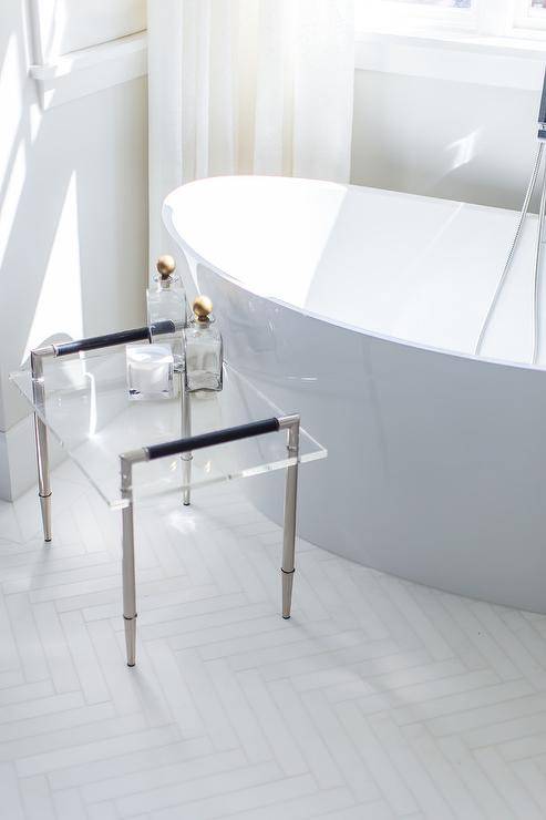 A nickel and lucite accent table sits on white herringbone floor tiles beside an oval freestanding bathtub positioned beneath a window dressed in white curtains.