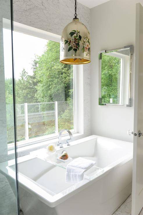 A gold lantern lights a rectangular freestanding bathtub fitted with a lucite bath tray and a chrome deck mount tub filler mounted in front of a picture window.