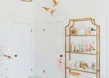 White nursery designed with a white herringbone Foraker Cow Hide Hand-Woven Cream Area Rug furnished with a gold etagere with glass shelves. Creative Co-Op Wash Metal and White Wood Beads Chandelier finish the room with an elegant gold flare.