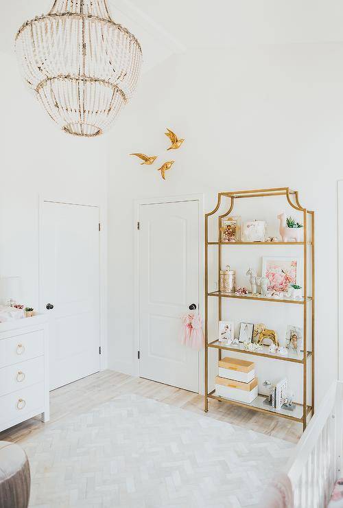 White nursery designed with a white herringbone Foraker Cow Hide Hand-Woven Cream Area Rug furnished with a gold etagere with glass shelves. Creative Co-Op Wash Metal and White Wood Beads Chandelier finish the room with an elegant gold flare.