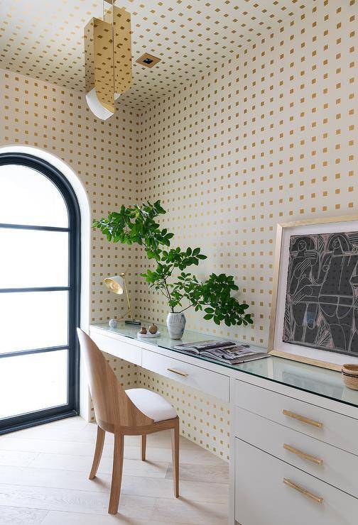 Clad in white and gold wallpaper, this welcoming home office features a walnut desk chair placed at a white built-in desk finished with a glass top.