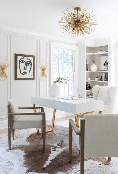 A brown cowhide rug is layered over a white and gray rug and placed in a white and gray home office boating a white wingback desk chair placed at a white and gold desk. The desk, illuminated by a brass sputnik flush mount, is paired with wooden chairs accented with gray cushions. Brass sconces, mounted to a wall finished with white moldings, flank an abstract art piece in a brown wooden frame, as a corner built-in bookcase is styled and complemented with gray grasscloth wallpaper on the backs of the shelves.