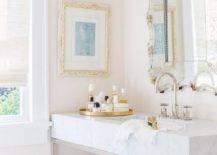 Lovely pale pink bathroom boasts an ornate vanity mirror hung above a nickel and marble washstand finished with a polished nickel gooseneck faucet. The vanity is topped with a round gold tray as a window is covered in a white linen roman shade.