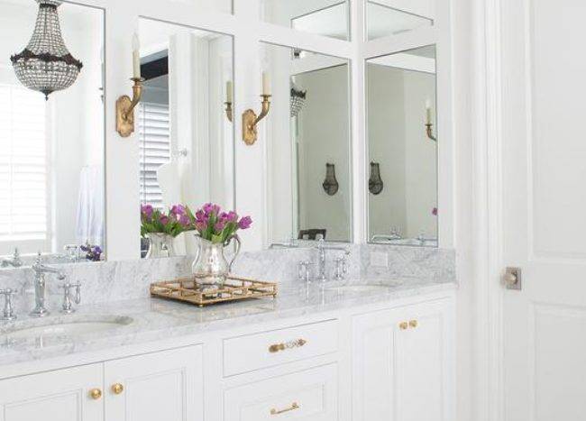 White bathroom with gold accents boasts a white dual vanity fitted with polished brass vanity knobs and a white marble countertop topped with a brass mirrored tray located beneath a wall lined with inset vanity mirrors lit by antique brass sconces.