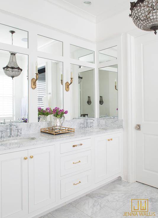 White bathroom with gold accents boasts a white dual vanity fitted with polished brass vanity knobs and a white marble countertop topped with a brass mirrored tray located beneath a wall lined with inset vanity mirrors lit by antique brass sconces.