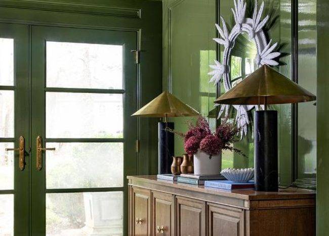 Contemporary foyer features a brown oak credenza lit by black marble lamps with brass shades on a glossy green lacquered wall with a white sunburst mirror, green French doors, green octagon ceiling molding and black and white harlequin floor tiles.