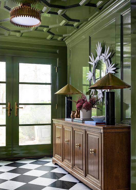 Contemporary foyer features a brown oak credenza lit by black marble lamps with brass shades on a glossy green lacquered wall with a white sunburst mirror, green French doors, green octagon ceiling molding and black and white harlequin floor tiles.