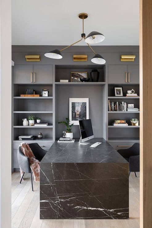 A honed black marble waterfall edge desk paired with charcoal gray chairs is fixed between built-in gray bookshelves lit by brass picture lights.