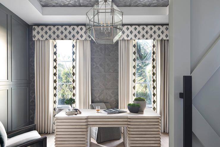 Elegant home office boasts a CFC Milo Desk paired with a gray slipper chair and positioned between windows covered in white curtains with a black diamond border and white and black diamond print valance. The window is framed by gray parquet wood wall tiles, while the room is lit by a Suzanne Kasler Morris Lantern hung from a parquet tiled gray ceiling.