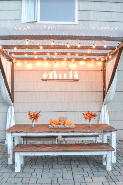 pergola hanging off side of house with string lights and candle chandelier hanging in middle modern picnic table underneath