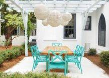 Wicker chandeliers hang from a white pergola over a teak dining table surrounded by aqua blue dining chairs.