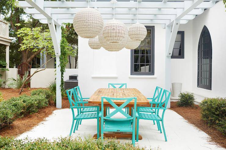 Wicker chandeliers hang from a white pergola over a teak dining table surrounded by aqua blue dining chairs.