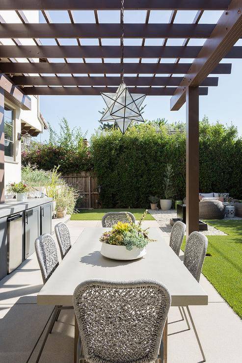 A Moravian star pendant hangs from a pergola over a white outdoor dining table surrounded by gray woven dining chairs.