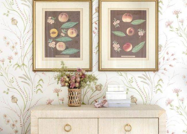 Vintage botanical prints in gold frames hang from a wall covered in Sanderson Summer Harvest wallpaper over a raffia wrapped dresser.