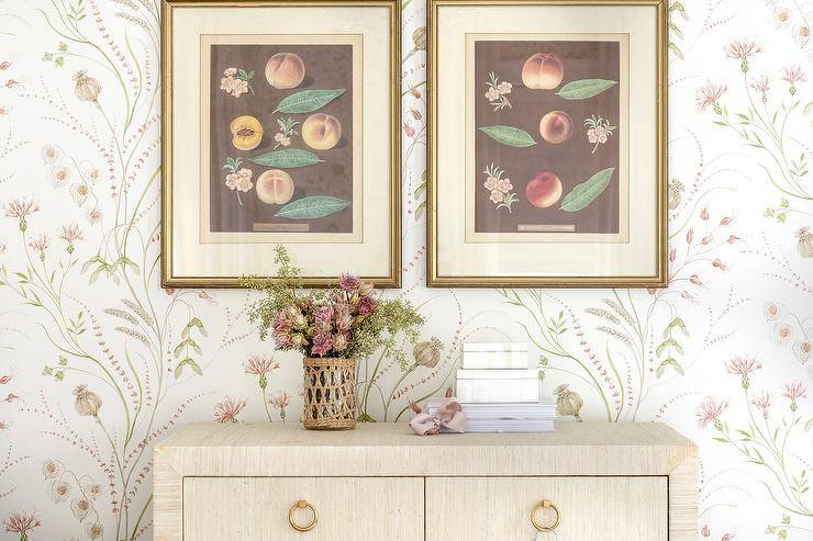 Vintage botanical prints in gold frames hang from a wall covered in Sanderson Summer Harvest wallpaper over a raffia wrapped dresser.