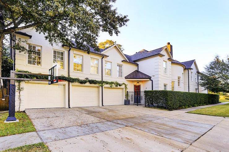 Gorgeous home boasts an ivory exterior alongside a gray roof over triple side by side garages framed by climbing plants. An iron gate opens up onto the front door entrance with stained wood door below a metal accent roof with clipped hedge highlighting the main facade which features a curved stairwell with staggered windows.