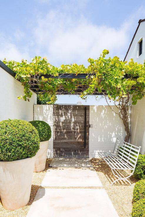 Large concrete pavers lead past a white metal bench to a gray plank wooden French door positioned beneath vines covering a black pergola.