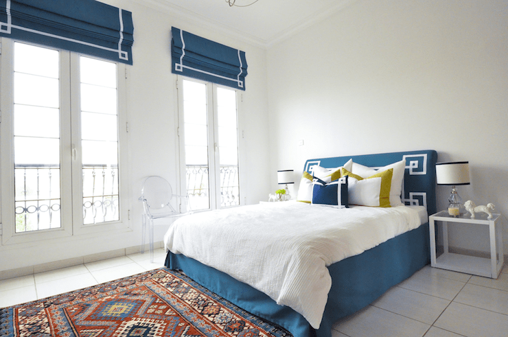 Gorgeous bedroom with vaulted ceilings and tiled floors. The bedroom features a peacock blue bed with upholstered headboard finished with white grosgrain trim. The bed is layered with white bed linens accented with mustard yellow bordered euro shams and a navy blue pillow with white grosgrain border. A white cube shaped glass topped nightstand flanks the bed topped with a glass double gourd table lamp with drum shade trimmed in navy. A Kartell Ghost Chair stands beside the bed between tall French doors leading to a Juliet balcony. A colorful Kilim style rug layers on the tiled floor at the foot of the bed. The tall French doors are dressed with peacock blue drapes with white grosgrain trim that echoes the detail of the headboard.