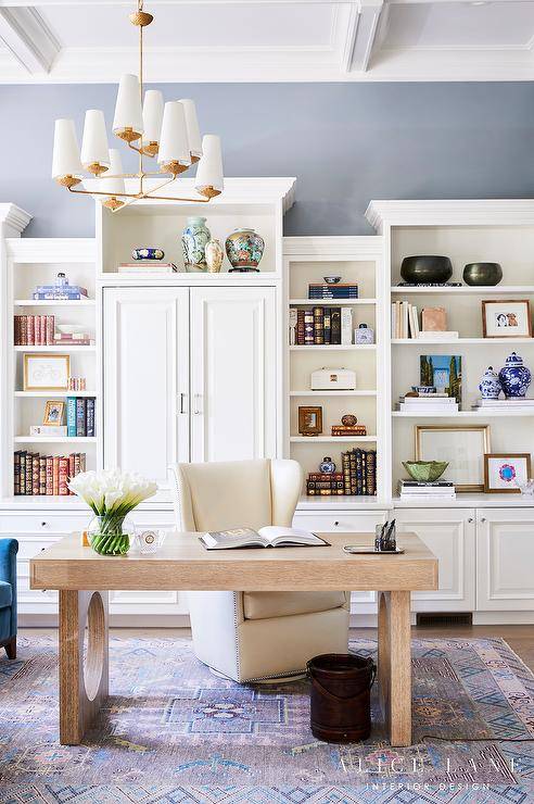 Home office design features an Aerin Fontaine large offset chandelier over a light brown oak desk with a cream wingback chair atop a blue and purple rug, white built in shelves and a white coffered ceiling.