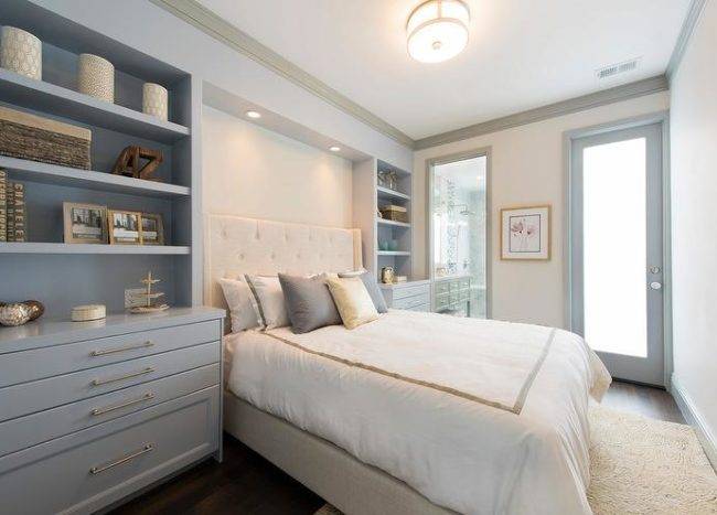 Placed in a headboard nook custom lit by recessed lighting, an ivory tufted wingback headboard supporting a bed dressed in white and gray bedding topped with layered gray and yellow pillows flanked by styled bluish gray built-in shelves positioned above gray drawers fitted with brass pulls. The bed sits on an ivory rug lit by a flush mount light as walls are framed by gray crown molding and baseboards.