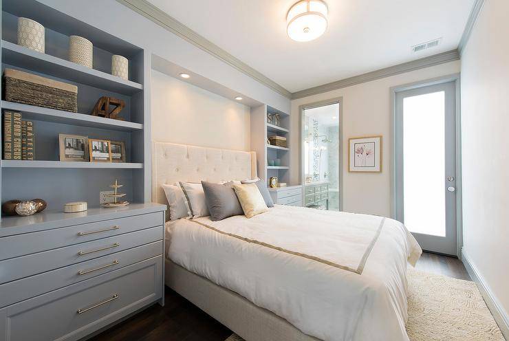 Placed in a headboard nook custom lit by recessed lighting, an ivory tufted wingback headboard supporting a bed dressed in white and gray bedding topped with layered gray and yellow pillows flanked by styled bluish gray built-in shelves positioned above gray drawers fitted with brass pulls. The bed sits on an ivory rug lit by a flush mount light as walls are framed by gray crown molding and baseboards.