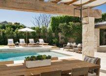 Stone pillars accent a rustic wood pergola finished with canvas and positioned over a teak plank dining table surrounded by teak slatted back dining chairs placed in front of an in ground swimming pool.