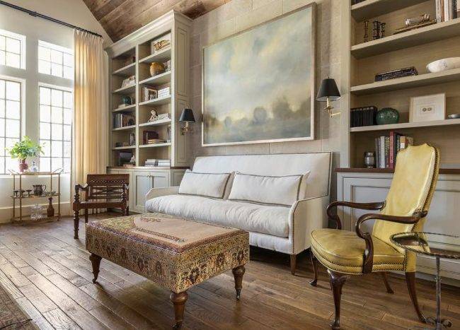 A large landscape art piece hangs over a white nailhead sofa placed facing a kilim bench and paired with a yellow leather French accent chair. The sofa is flanked by light gray built-in shelves mounted over light gray cabinets.